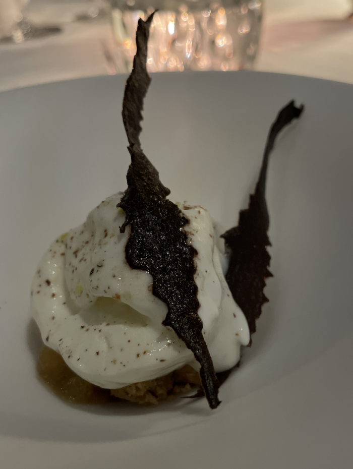 In a white bowl, a white scoop of ice cream with dark flecks, topped with what looks like dark leaves, but they're chocolate